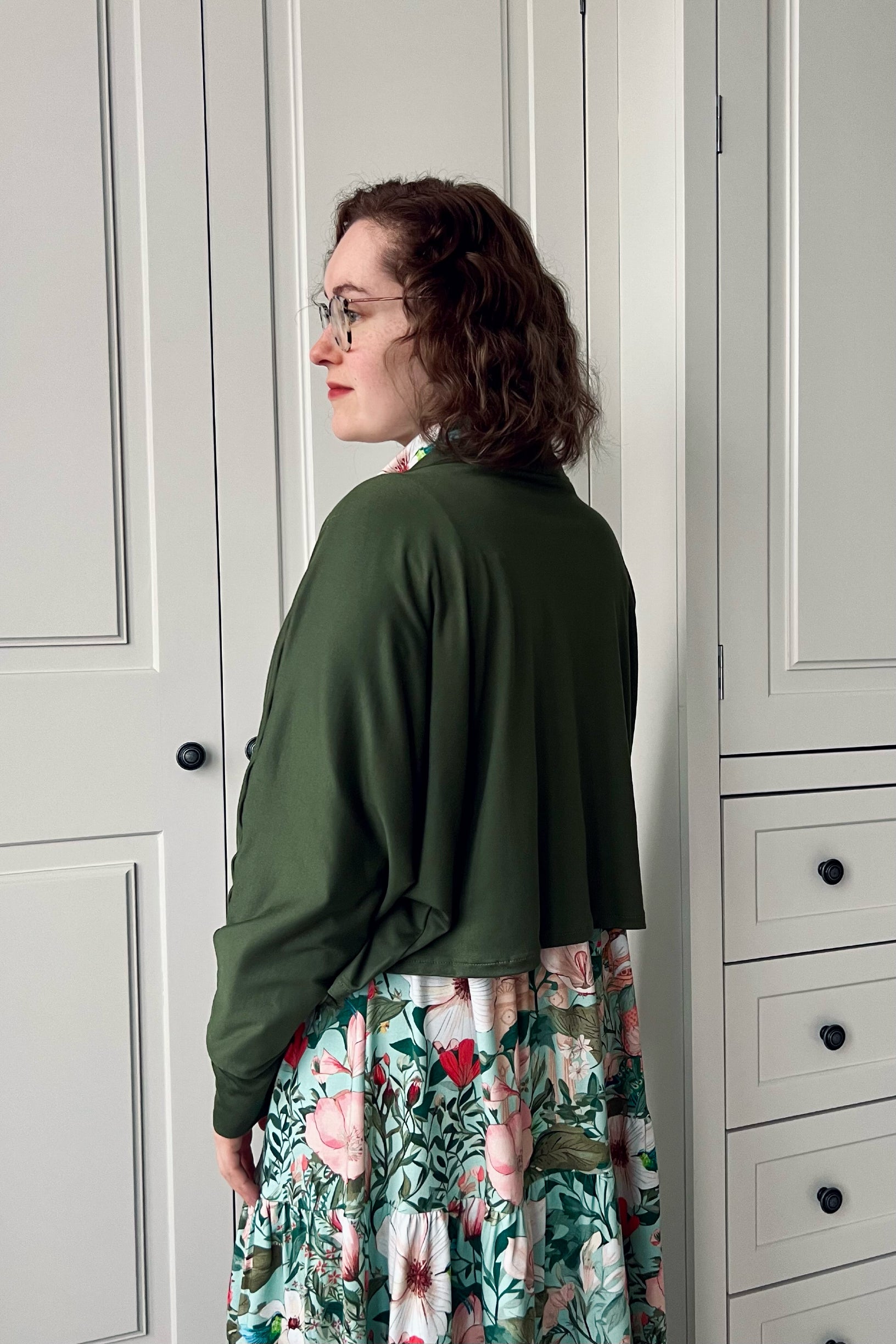 Woman wearing a green batwing cardigan and floral dress standing in front of a wardrobe.