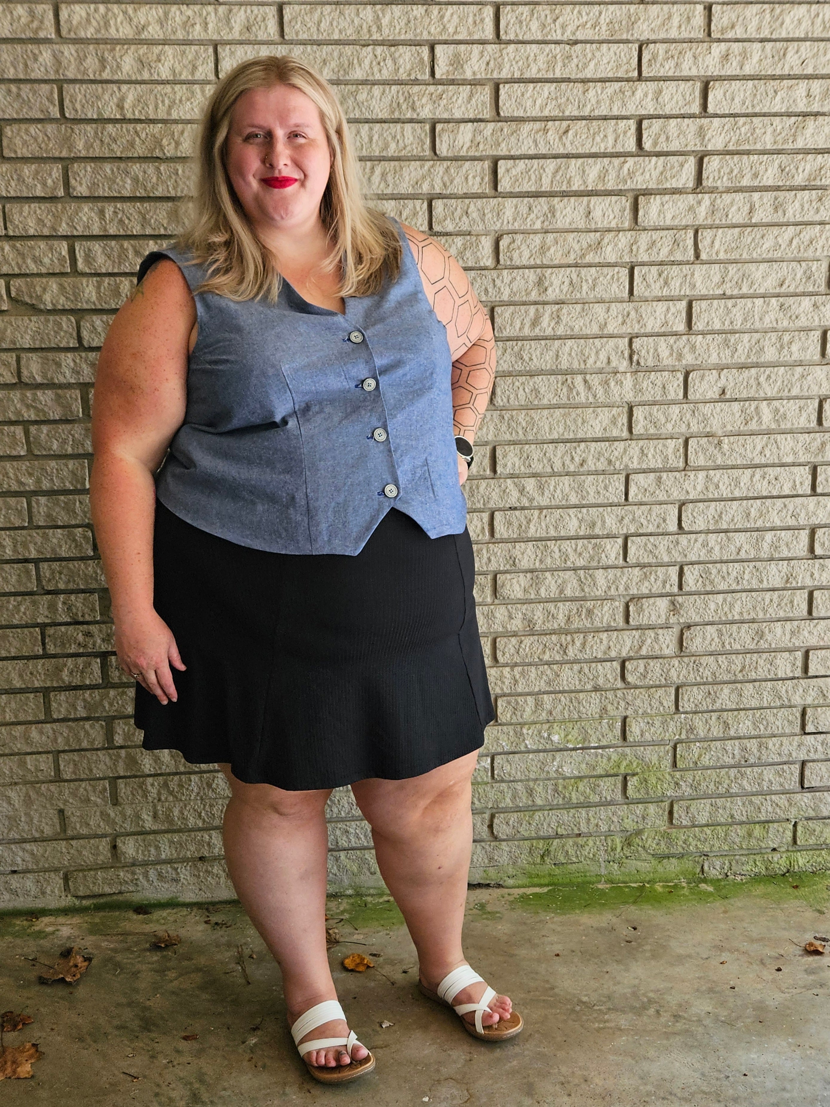 Woman wearing a denim vest over a black dress against a brick wall.