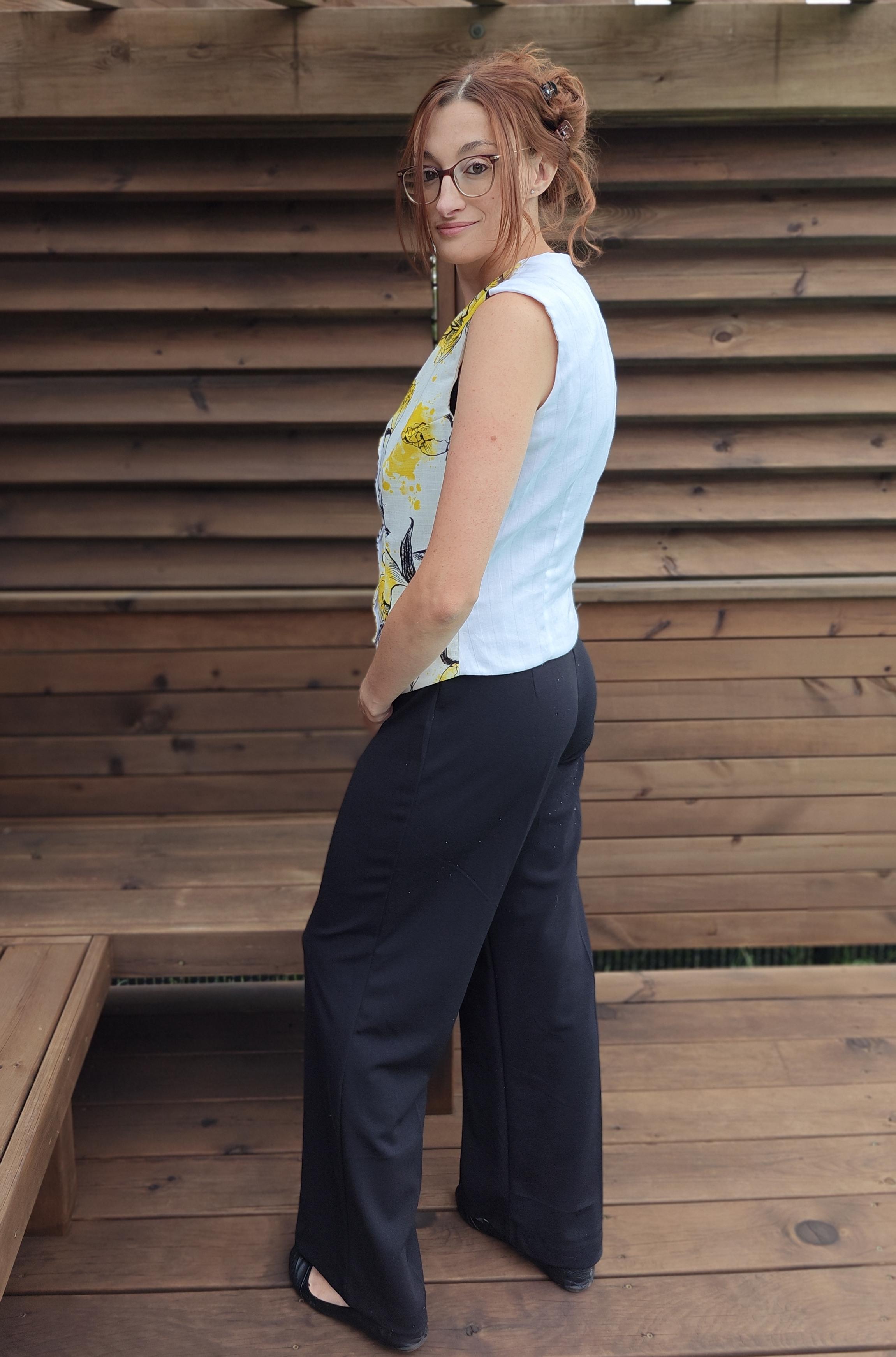 Woman wearing a floral top and black pants standing against a wooden wall.