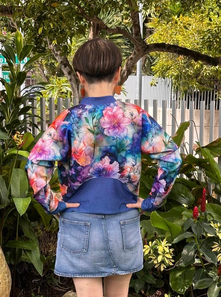 Woman outside modeling a floral and blue athletic bomber jacket with zip closure, kangaroo pocket detail, and cuffs. Easy pdf sewing pattern.