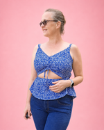 A woman modeling a blue and white patterned cutout top made using Ellie and Mac Sewing patterns with blue jeans, standing against a solid pink background.