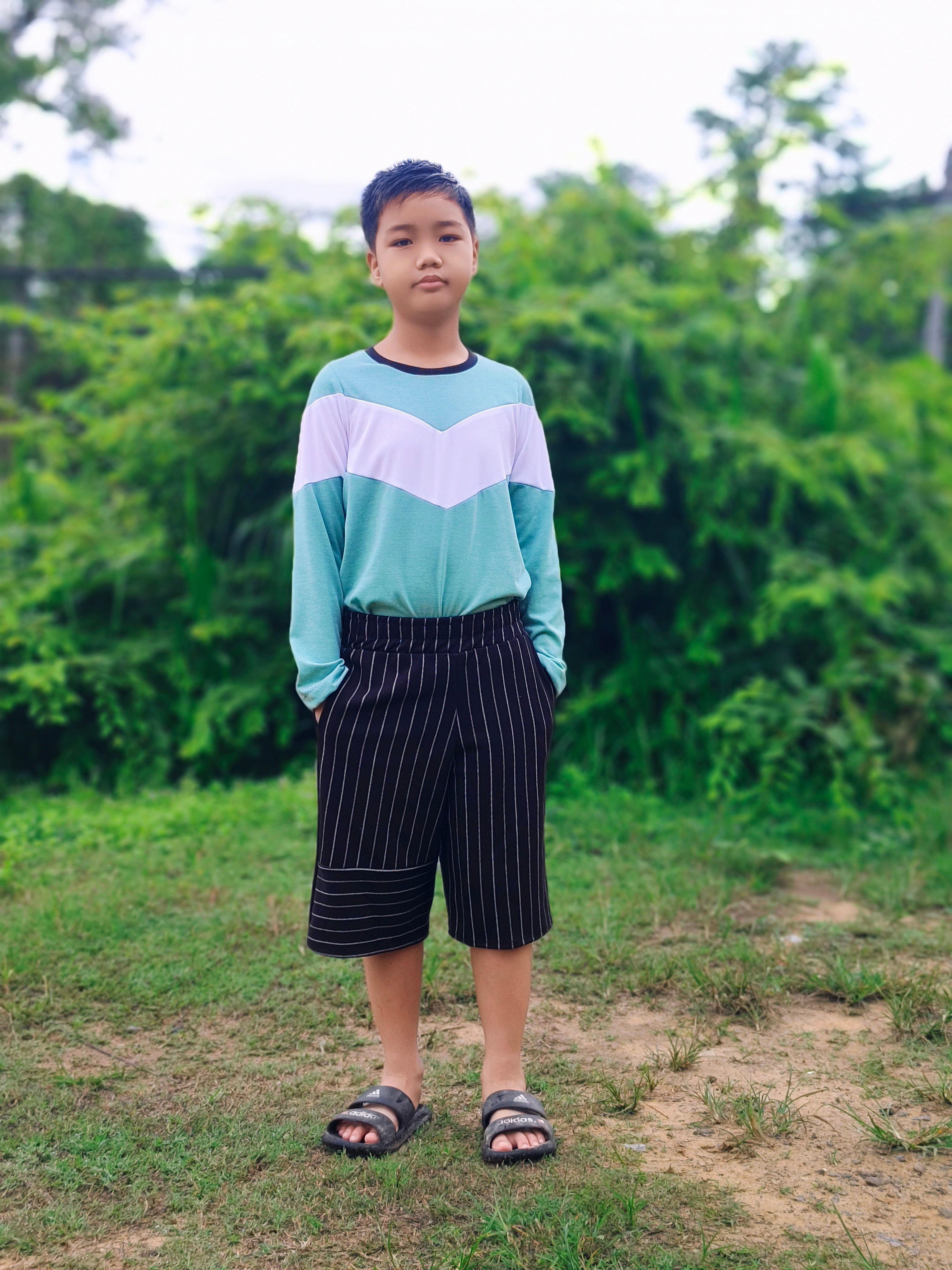 Person standing outdoors on a grassy area with trees in the background wearing long striped shorts.