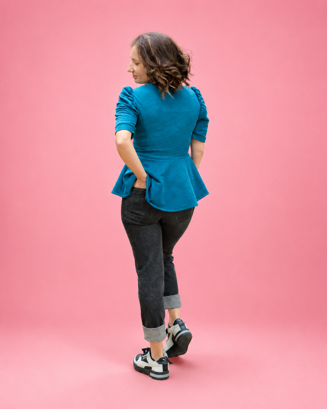 A woman is shown from behind wearing a bright teal blue peplum top, dark gray rolled-calf jeans, and black and white sneakers, standing against a solid pink studio background. The top is made using Ellie and Mac PDF sewing patterns. They include A4, Letter, A0, and Projector pattern files. They also include a full sew-along tutorial. 