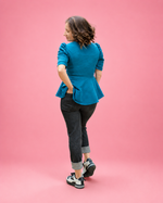 A woman is shown from behind wearing a bright teal blue peplum top, dark gray rolled-calf jeans, and black and white sneakers, standing against a solid pink studio background. The top is made using Ellie and Mac PDF sewing patterns. They include A4, Letter, A0, and Projector pattern files. They also include a full sew-along tutorial. 