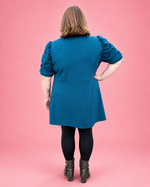 A woman viewed from the back wearing a teal blue puff-sleeve tunic dress with black tights and brown boots, standing against a solid pink studio background.