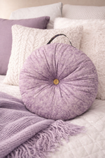 Back view of a Round purple pillow with a button center and handle on a bed with white and purple bedding. Made using Ellie and Mac sewing patterns.