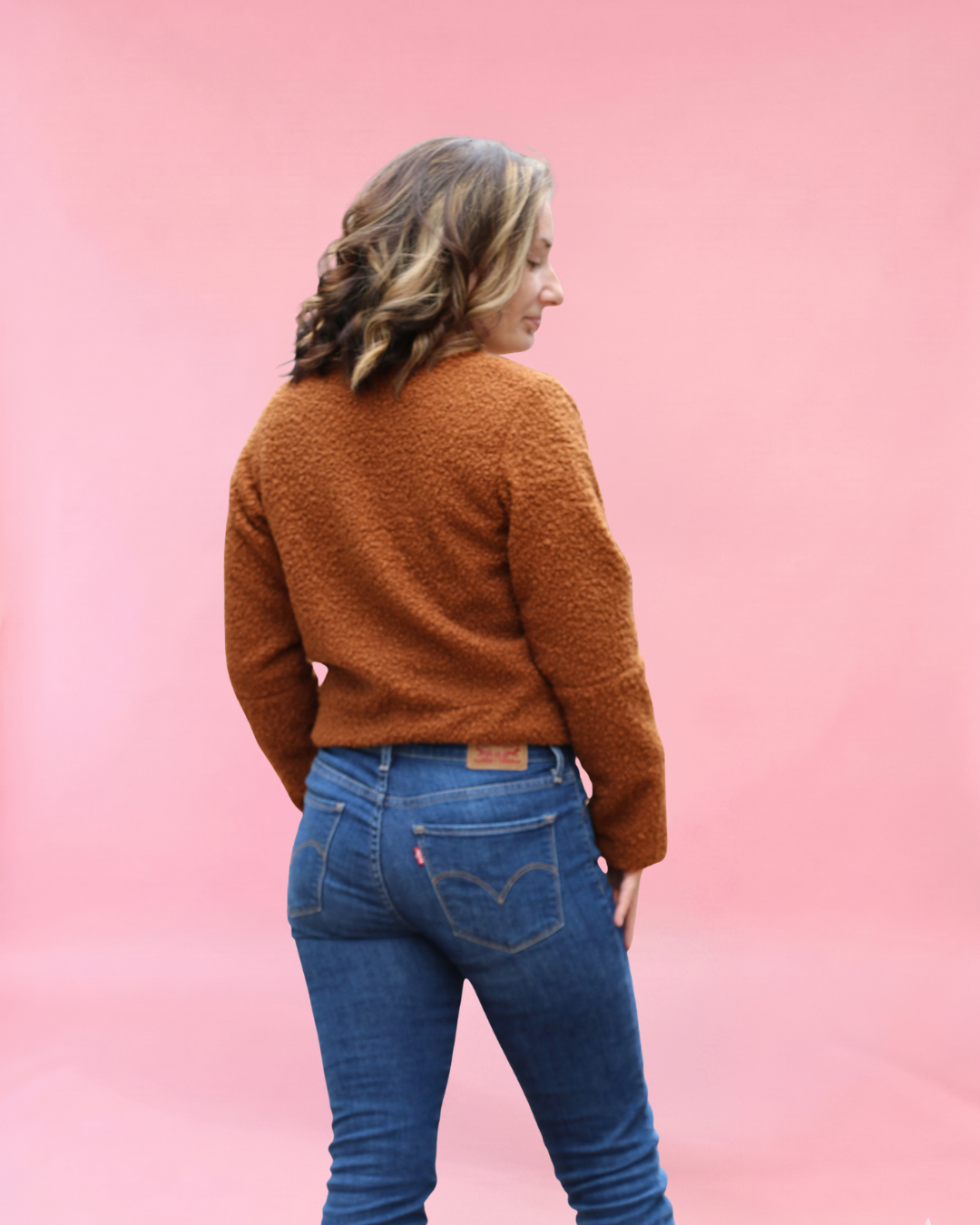Person wearing a brown sherpa jacket and blue jeans against a pink background