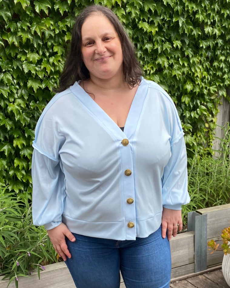 Person wearing a light blue cardigan with gold buttons and blue jeans standing outdoors.