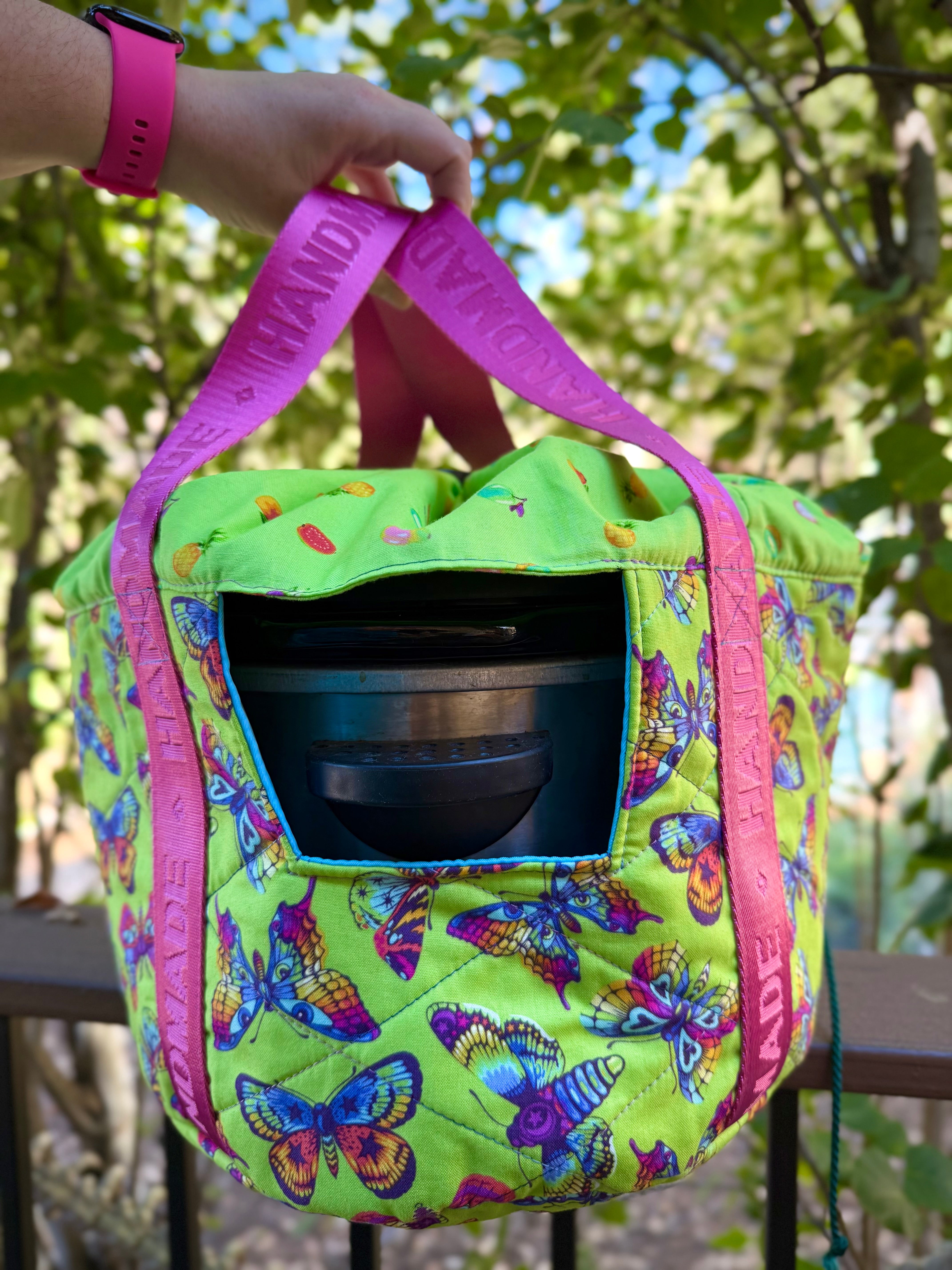 Green crock pot carrier bag with butterfly pattern and pink straps holding a black object, outdoors. Shows the hole for the handles for easy carrying.