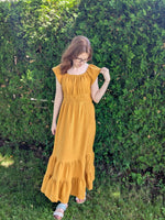 Woman wearing a long yellow dress standing in front of green foliage. The dress is tiered and has cap sleeves. Easy boho style. Womens sewing patterns.
