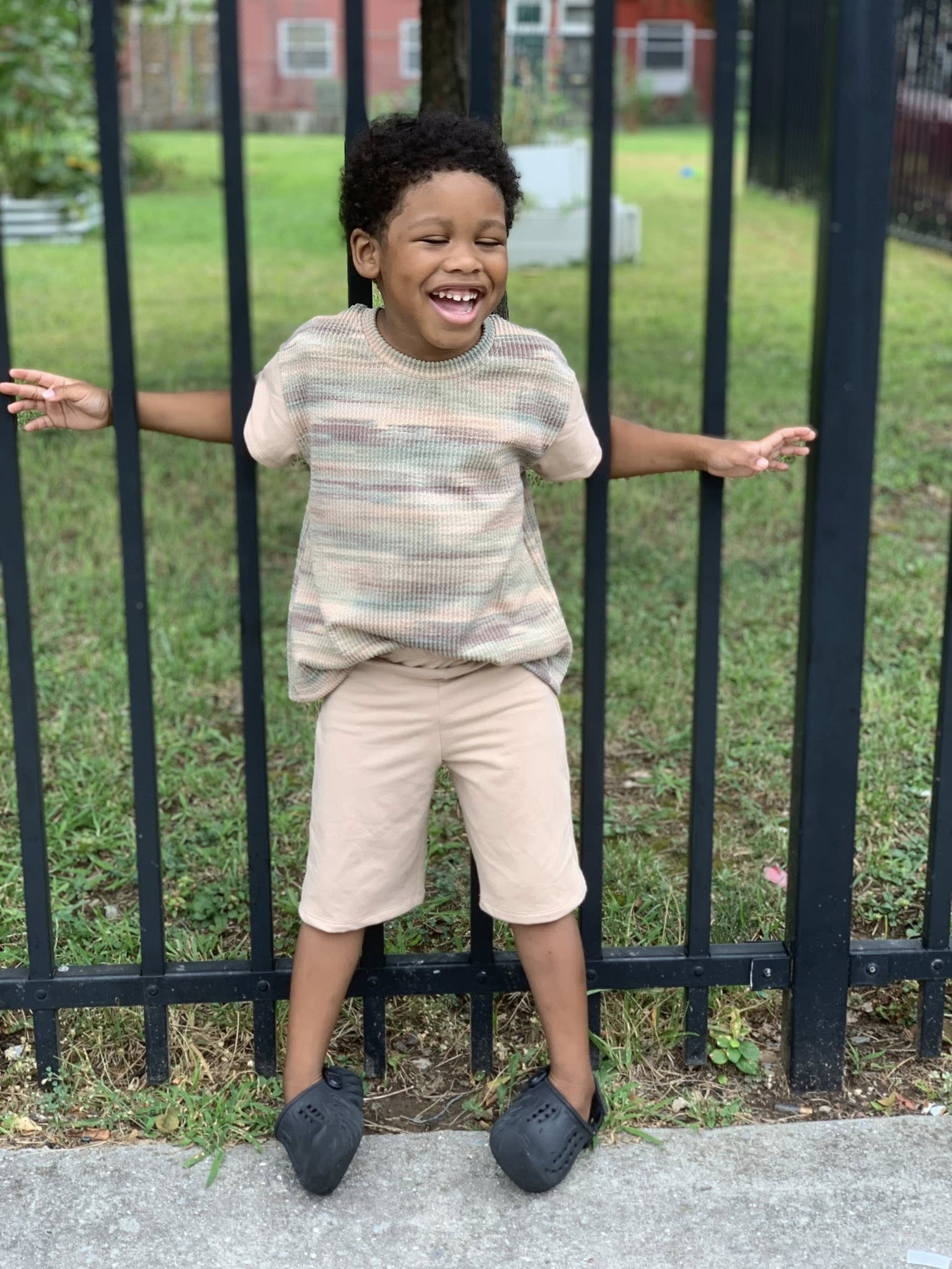 Child standing with arms outstretched in front of a black metal fence with grass and houses in the background.