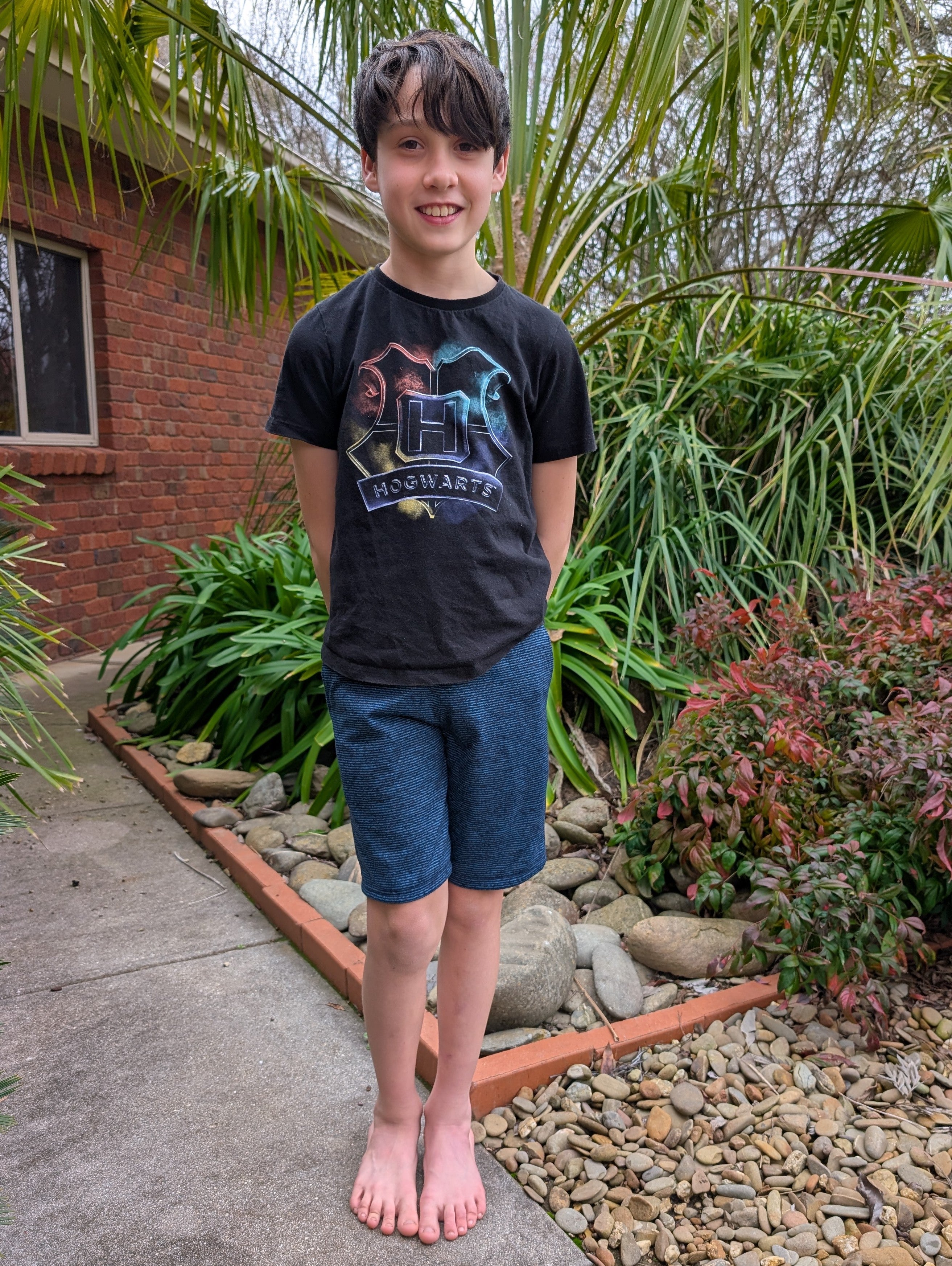 Young boy standing outdoors in a garden with plants and rocks around him. Wearing blue athletic shorts and a graphic tee. 