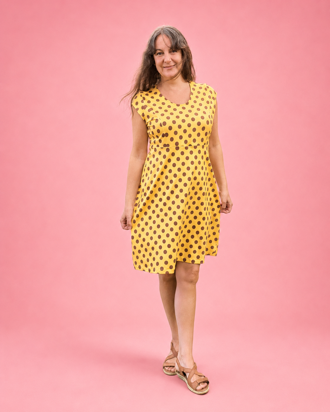 A woman modeling a yellow sleeveless V-neck dress with black polka dots, standing against a solid pink studio background.
