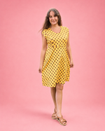 A woman modeling a yellow sleeveless V-neck dress with black polka dots, standing against a solid pink studio background.