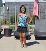 Ellie & Mac Gathered Tankini Swimsuit Sewing Pattern. The image shows a woman modeling the finished swimsuit in a vibrant abstract print, alongside a pool. This PDF pattern is available in sizes XXS-6XL and printable on A4, A0, and Letter paper sizes. Projector file and video tutorial included. 