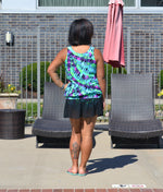 Ellie & Mac Gathered Tankini Swimsuit Sewing Pattern. The image shows a woman modeling the finished swimsuit in a vibrant abstract print, alongside a pool. This PDF pattern is available in sizes XXS-6XL and printable on A4, A0, and Letter paper sizes. Projector file and video tutorial included. 