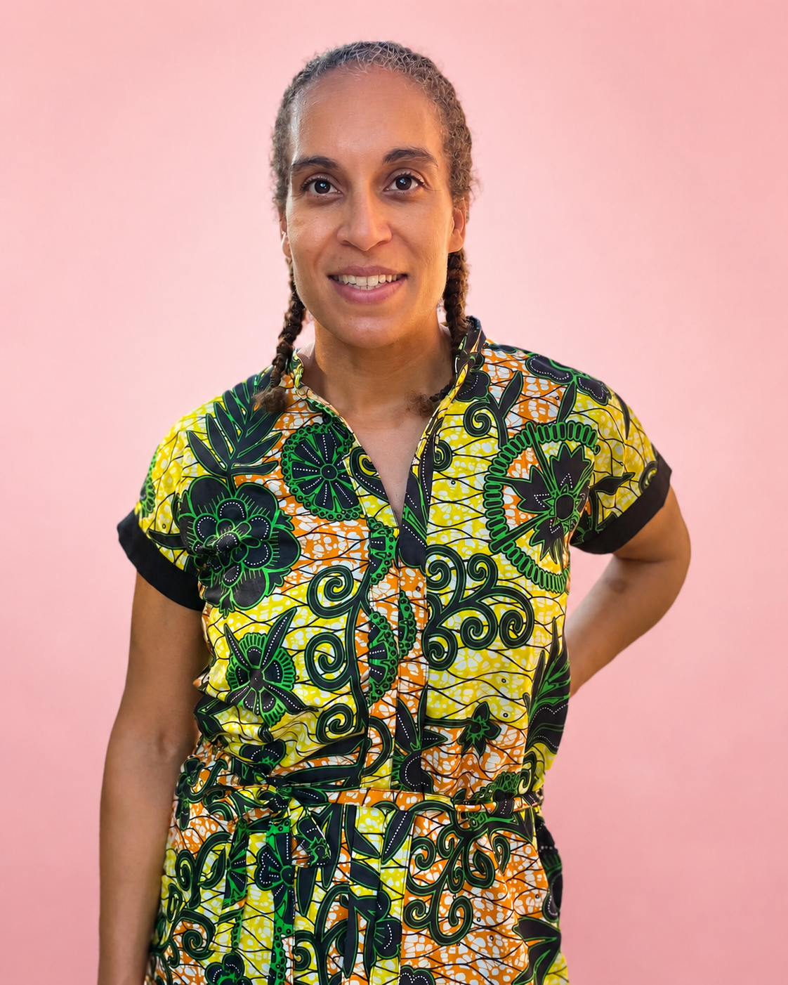 A woman with braided hair wearing a short-sleeve button-up shirt with a vibrant yellow, green, and black batik-style floral pattern, posed against a solid pink background. Jade dress pattern.