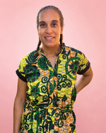 A woman with braided hair wearing a short-sleeve button-up shirt with a vibrant yellow, green, and black batik-style floral pattern, posed against a solid pink background. Jade dress pattern.
