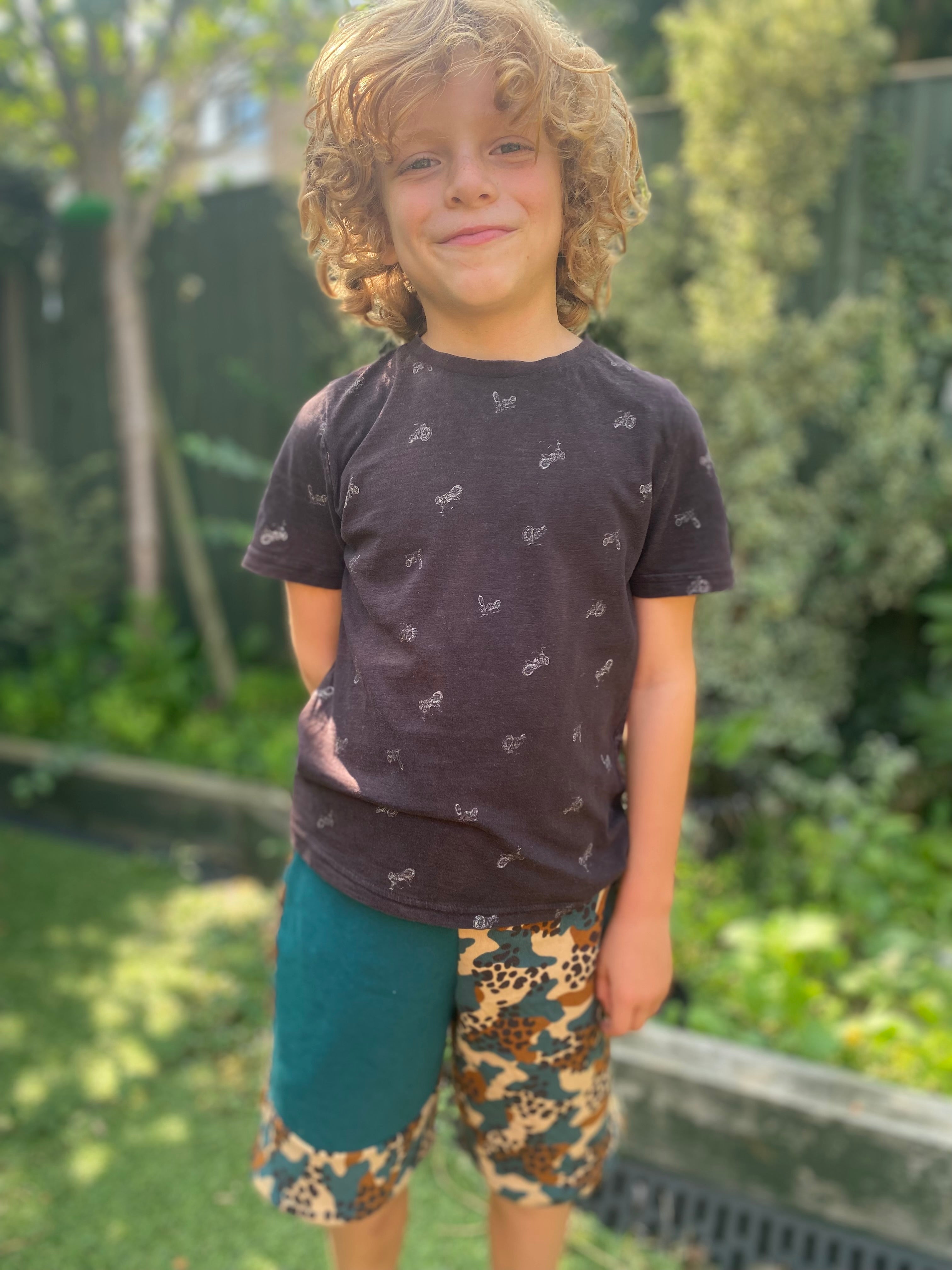 Child wearing a dark t-shirt with small patterns and colorful shorts standing outdoors.
