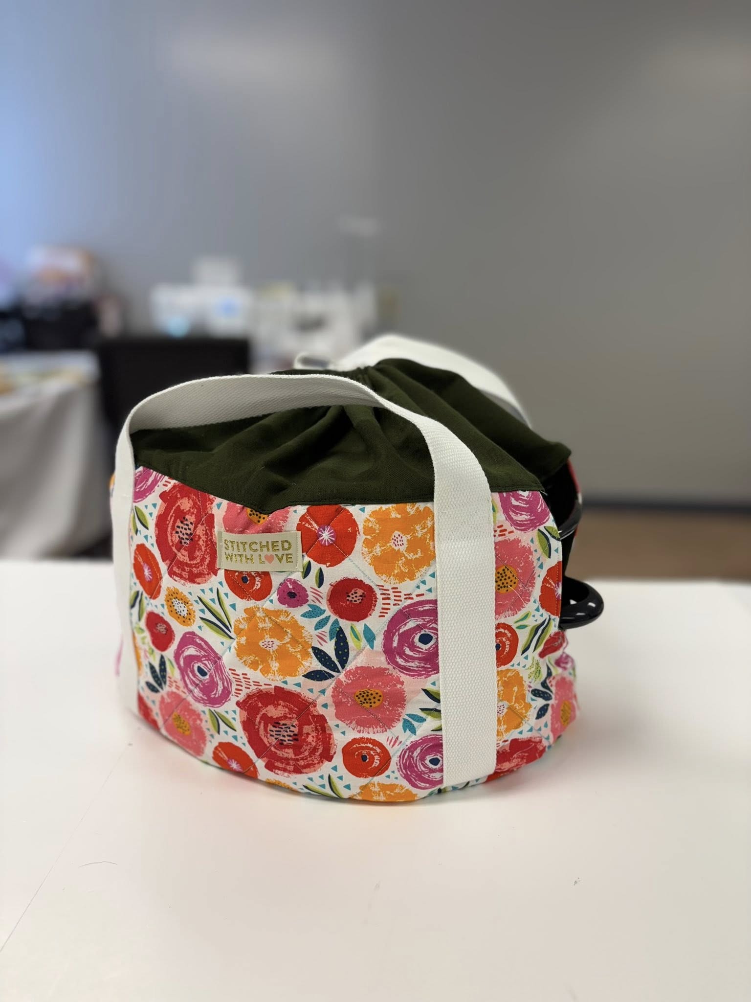 Floral-patterned slow cooker carrying bag with white straps and green top on a white surface with a blurred background. Easy home craft sewing patterns by Ellie and Mac Patterns.