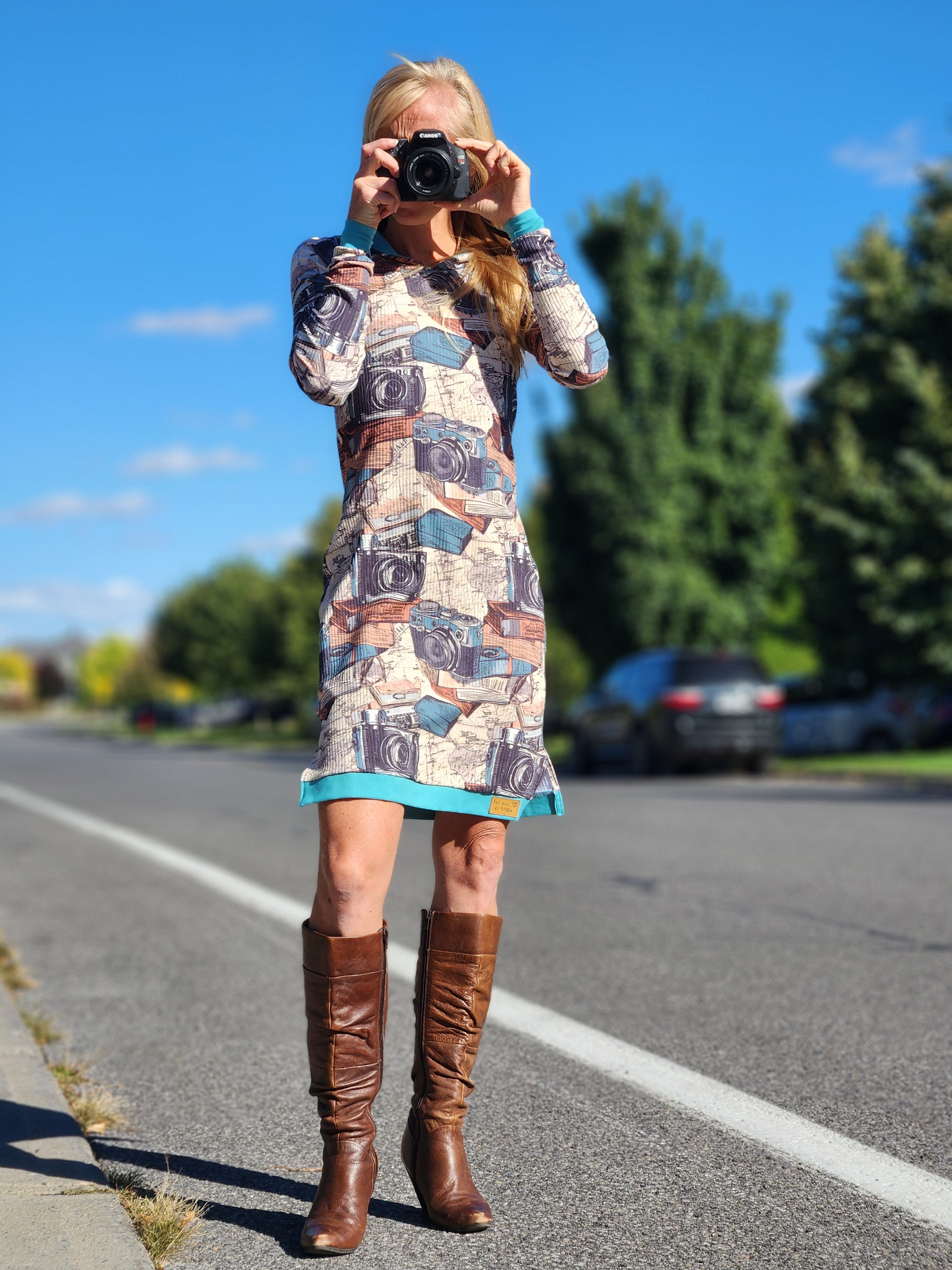 Person wearing a camera print fabric dress taking a photo on a road with trees in the background