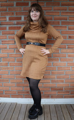 Woman wearing a brown dress with a belt against a brick wall.