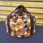 Multicolored geometric-patterned slow cooker carrier bag with a brown strap against a striped background.