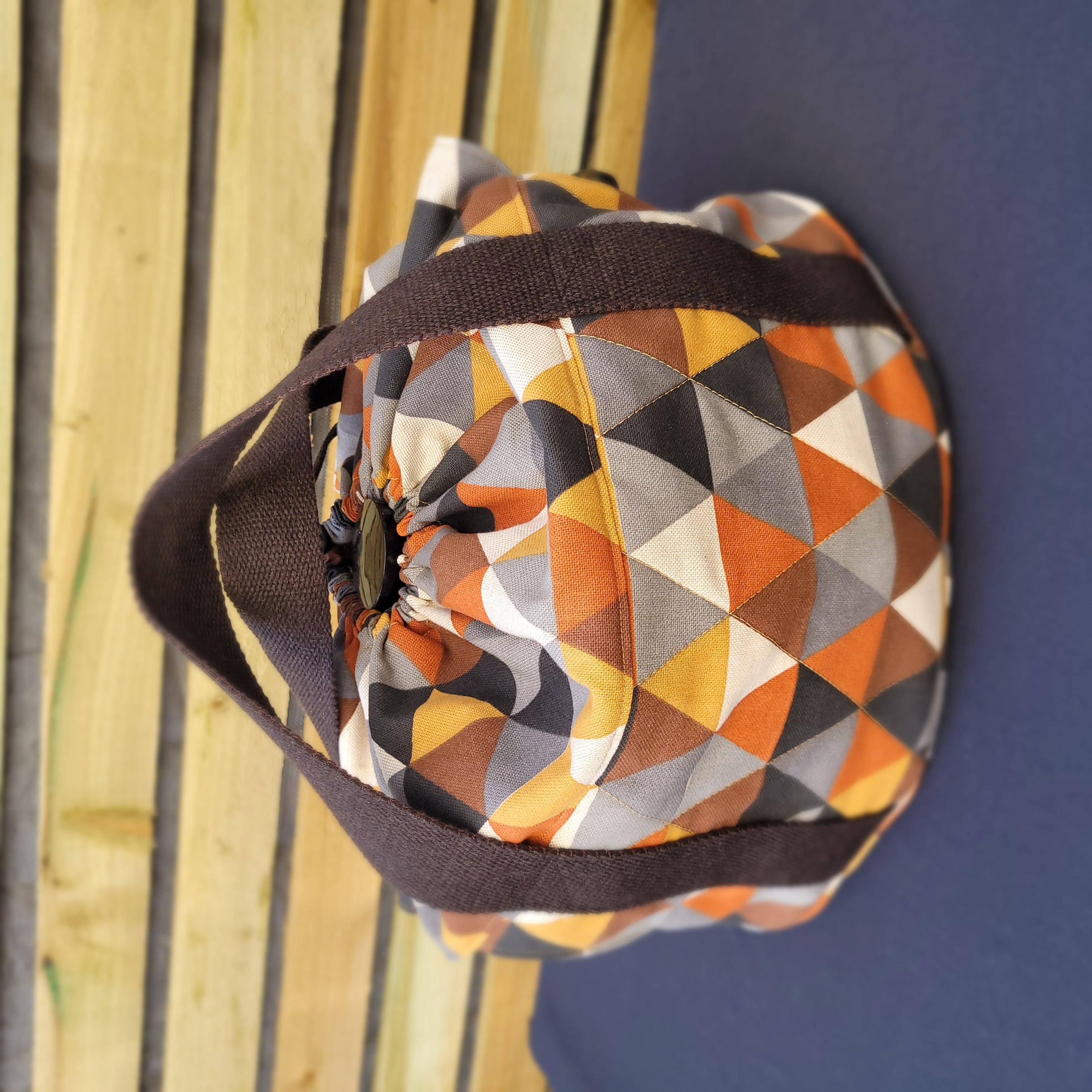 Multicolored geometric-patterned slow cooker carrier bag with a brown strap against a striped background.
