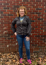 Person wearing a dark gray cardigan  with bishop sleeves over a graphic t-shirt, standing against a brick wall with fallen leaves on the ground.