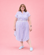 A woman modeling a light purple short-sleeve midi shirt dress with a white floral pattern, a white belt, and white slip-on shoes, standing against a solid pink background. She made this dress using Ellie and Mac PDF sewing patterns. They include A0, A4, Letter, print files and projector sewing file. They also include a full sew along video tutorial.
