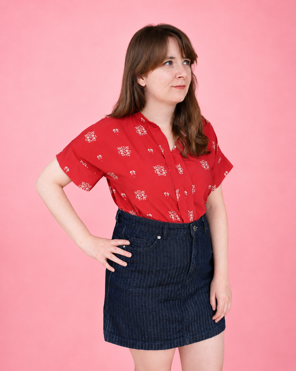 A woman modeling a woven red short-sleeve placket shirt with a white floral pattern, paired with a dark blue denim skirt, set against a solid pink background. Blouse sewn using using Ellie and Mac PDF sewing patterns. They include A0, A4, Letter, print files and projector sewing file. They also include a full sew along video tutorial.