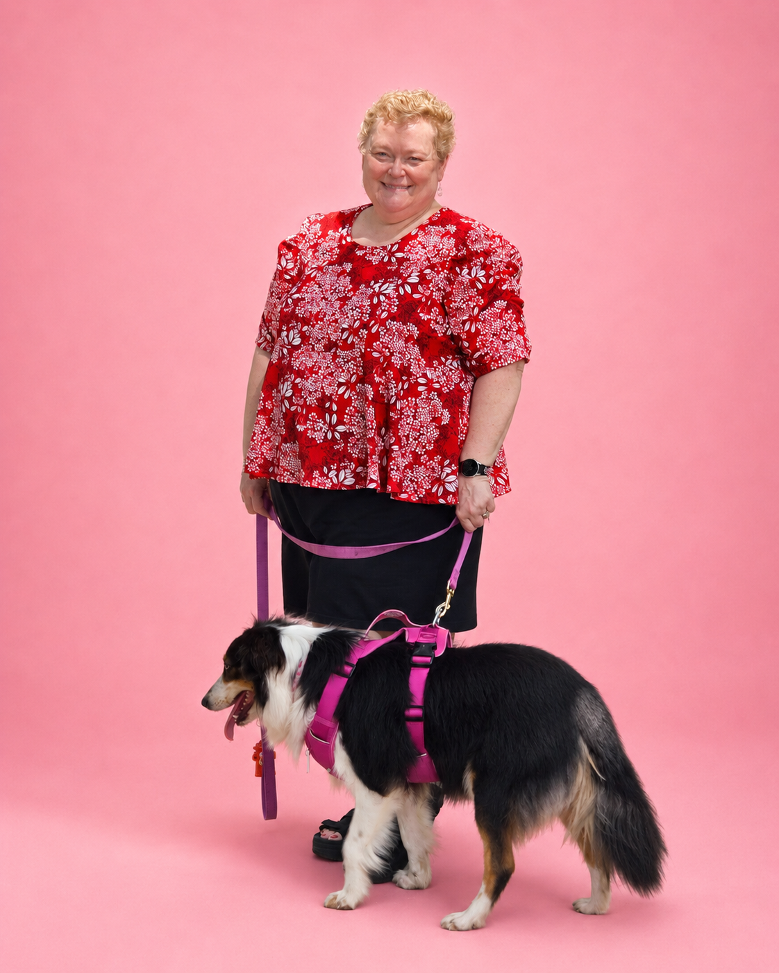 A woman stands on a pink studio background holding a black, white, and tan dog wearing a bright pink harness and leash. The woman wears a red floral short-sleeve top and a dark skirt. The top is made using Ellie and Mac PDF sewing patterns. They include A4, Letter, A0, and Projector pattern files. They also include a full sew-along tutorial. 