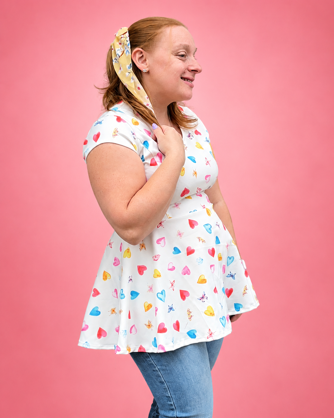 A woman wearing a white short-sleeve skater dress with a colorful heart and star pattern, paired with blue jeans and a matching patterned headband, posing against a solid pink background. The top is made using Ellie and Mac PDF sewing patterns. They include A4, Letter, A0, and Projector pattern files. They also include a full sew-along tutorial. 