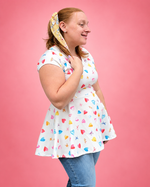 A woman wearing a white short-sleeve skater dress with a colorful heart and star pattern, paired with blue jeans and a matching patterned headband, posing against a solid pink background. The top is made using Ellie and Mac PDF sewing patterns. They include A4, Letter, A0, and Projector pattern files. They also include a full sew-along tutorial. 