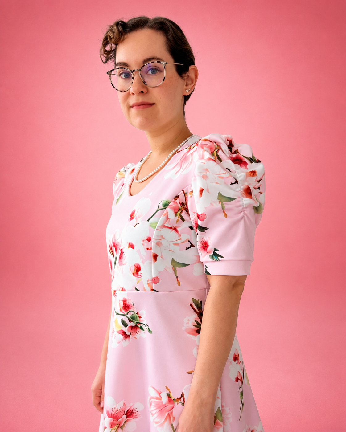 A woman with short dark hair and glasses wearing a light pink puff-sleeve dress with a red and green floral print, posed against a solid pink background. The dress is made using Ellie and Mac PDF sewing patterns. They include A4, Letter, A0, and Projector pattern files. They also include a full sew-along tutorial. 
