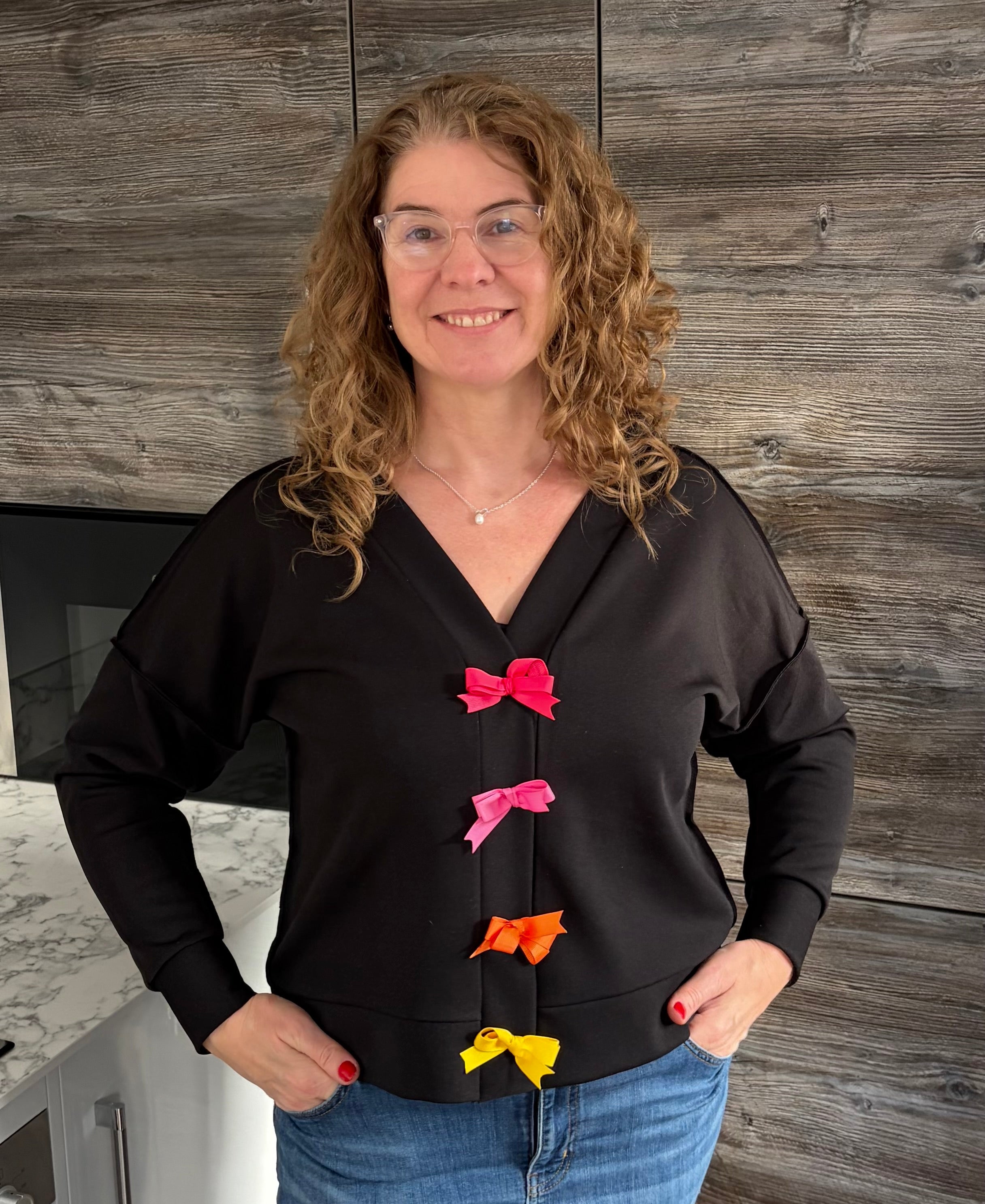 Woman wearing a black cropped cardigan sweater with colorful bows standing in a room with wooden walls.
