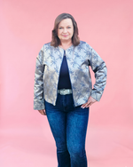 Woman wearing a patterned jacket and blue jeans standing against a wooden wall.