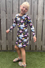 Child wearing a colorful dress with animal pattern standing in front of a wooden fence.