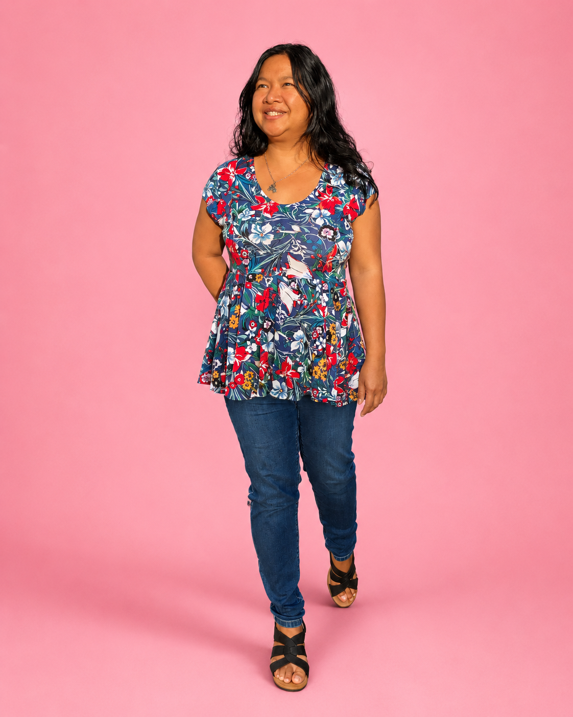 A woman modeling a short-sleeve peplum top with a colorful floral and bird pattern, paired with blue jeans and black sandals, standing against a solid pink background. The top is made using Ellie and Mac PDF sewing patterns. They include A4, Letter, A0, and Projector pattern files. They also include a full sew-along tutorial. 