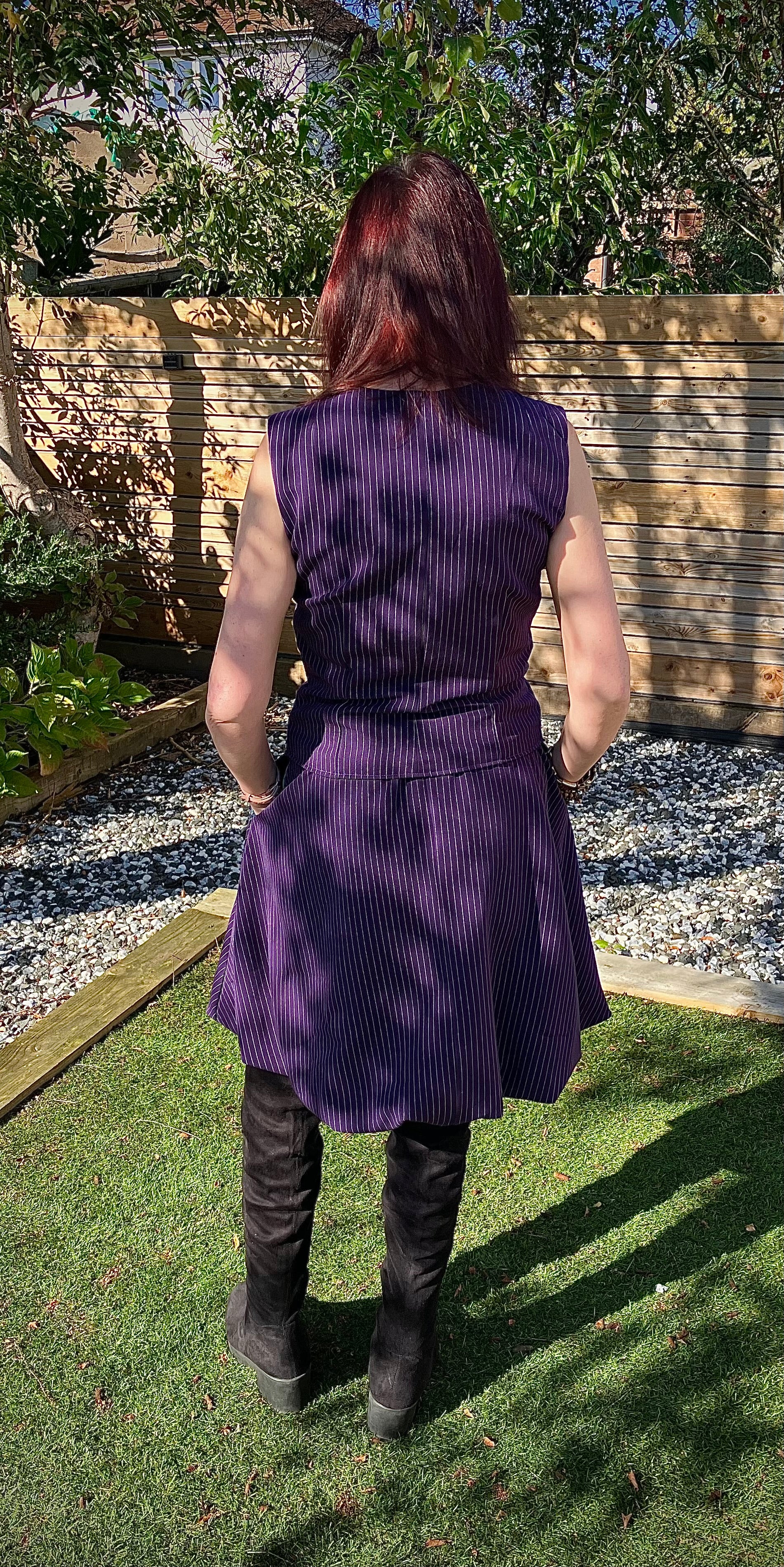 Person wearing a purple sleeveless bubble skirt, waistcoat, and black boots standing in a garden. This entire outfit was made using Ellie and Mac's Beginner Friendly sewing patterns. 