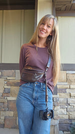 Woman in mauve long sleeve shirt and jeans, holding a camera that is attached to the camera strap that is an accessory to a brown leather belt bag the woman is wearing. Make your own belt bag with Ellie and Mac sewing patterns.