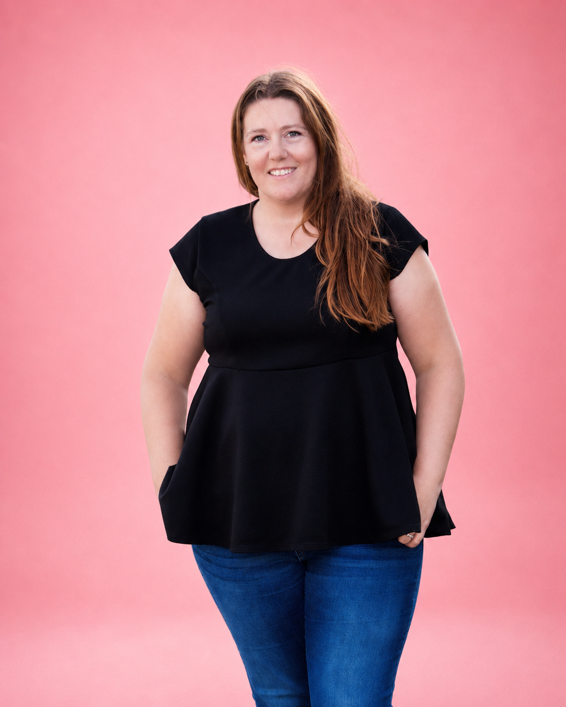 A woman with long light brown hair stands against a solid pink studio background, wearing a black short-sleeve peplum top and blue jeans. 