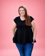 A woman with long light brown hair stands against a solid pink studio background, wearing a black short-sleeve peplum top and blue jeans. 