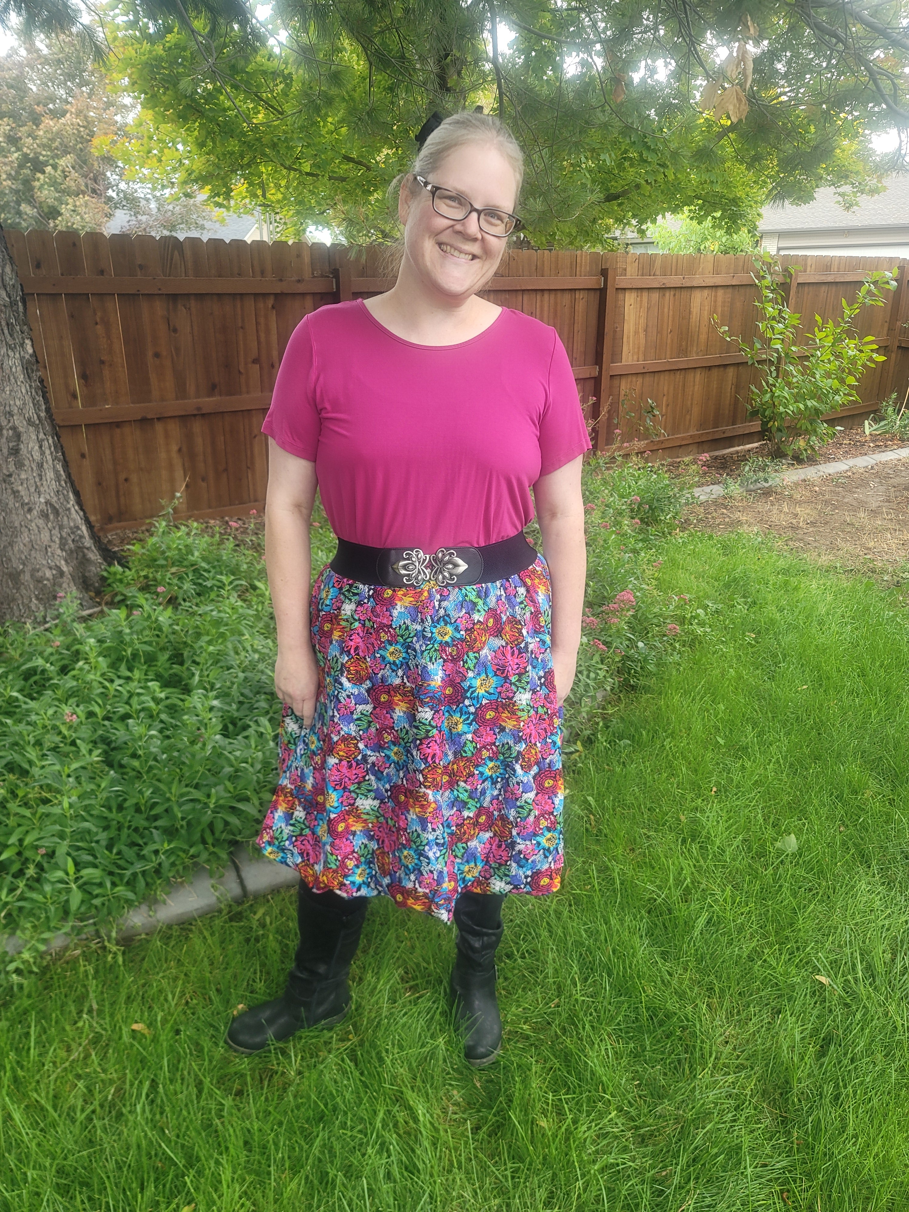 Person wearing a colorful floral dress and pink top standing on grass with a wooden structure and trees in the background.