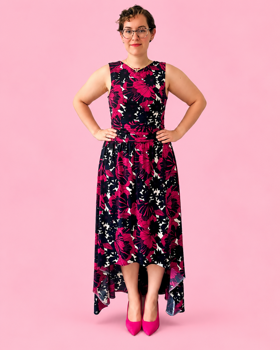 A woman modeling a sleeveless high-low dress with a bold floral pattern in black, magenta, and white, paired with bright pink heels, set against a solid pink background.