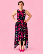 A woman modeling a sleeveless high-low dress with a bold floral pattern in black, magenta, and white, paired with bright pink heels, set against a solid pink background.