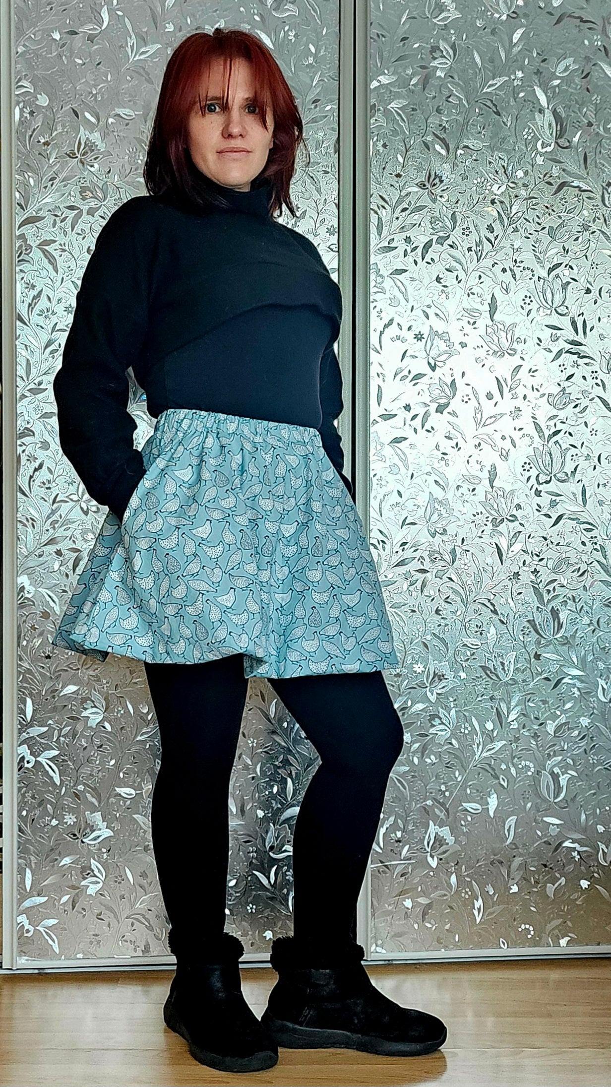 Person wearing a black turtleneck bolero, blue patterned bubble skirt, and black shoes standing in front of a frosted glass door. Outfit sewn using Ellie and Mac sewing patterns.