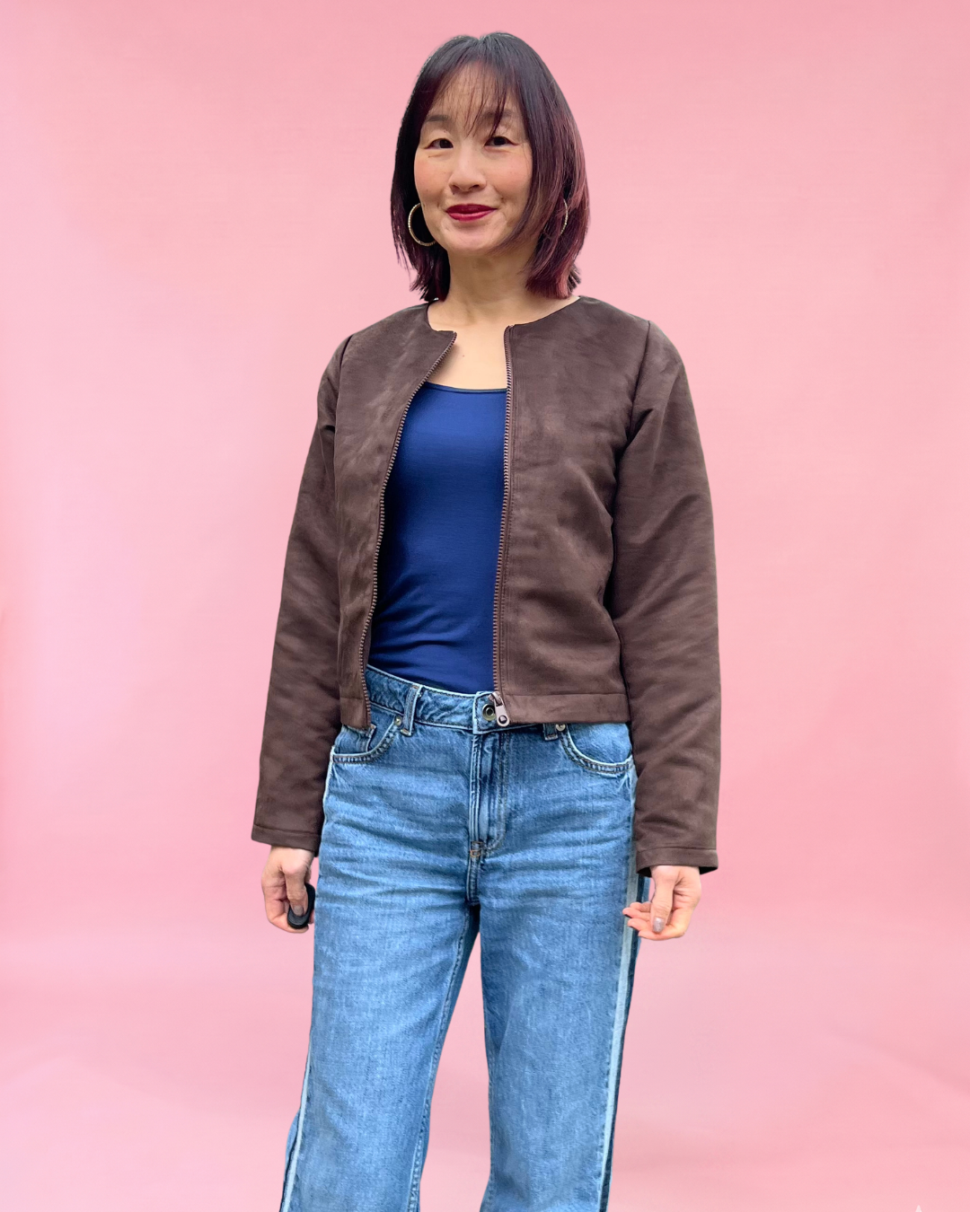 Woman wearing a brown suede jacket made using Ellie & Mac PDF sewing pattern and blue jeans against a pink background