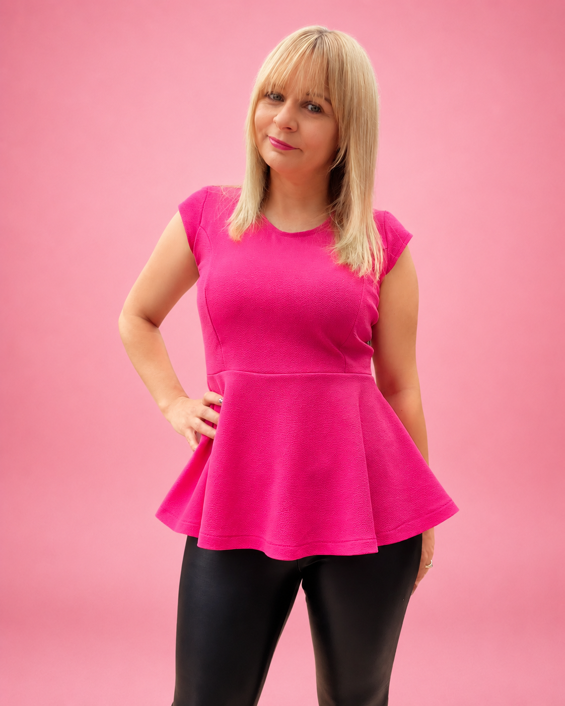A woman modeling a bright pink short-sleeve peplum top with black leggings, standing against a solid pink studio background. The top is made using Ellie and Mac PDF sewing patterns. They include A4, Letter, A0, and Projector pattern files. They also include a full sew-along tutorial. 