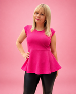 A woman modeling a bright pink short-sleeve peplum top with black leggings, standing against a solid pink studio background. The top is made using Ellie and Mac PDF sewing patterns. They include A4, Letter, A0, and Projector pattern files. They also include a full sew-along tutorial. 